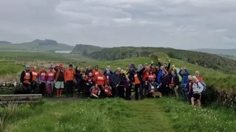 A group of walkers stand at Steelrigg near a stretch of Hadrian's Wall in Northumberland. They are mostly wearing orange t-shirts which have the logo of the Motor Neuron Disease Association.