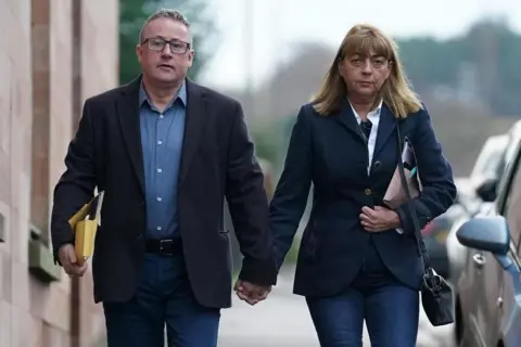 PA Media Stuart and Linda Allen walk down the street towards the camera hand in hand. He is wearing blue jeans, a blue shirt and a dark jacket and she is wearing blue jeans and a blue jacket over a white shirt. She has a handbag on her shoulder and both of them are carrying folders. 