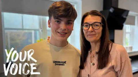 BBC A young man with short dark hair in a cream sweatshirt next to his mother with long dark hair, glasses and a pink cardigan. They are standing in a kitchen. With BBC Your Voice branding in white font in the bottom left of the image