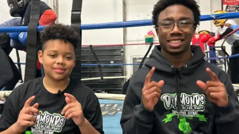 Two young boxers smile and point to the camera. They are wearing black hoodies that say Diamonds on the Ruff Boxing Gym. They are sitting on the edge of a boxing ring where two boxers are sparring.