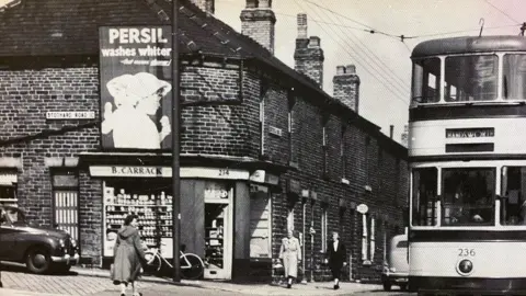Carracks A historical black and white street scene showing a terraced building with tall chimneys. There is a sign on a shop front which reads 'B Carrack'. There are three people walking and a double decker bus. 