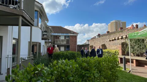 White two-floor apartments on the left with balconies. There is green hedges at the centre and people in the distance talking with one another. Behind the people is a building with more balconies and another large building to the left. 