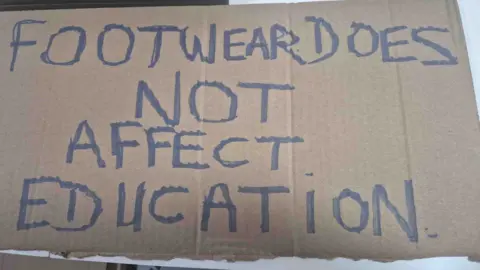 Duaine Davies Cardboard sign reading: 'Footwear does not affect education'.