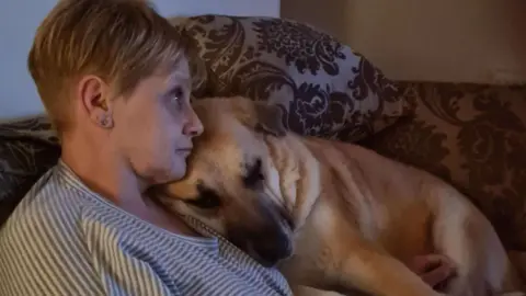 Caroline Dewson Caroline Dewson, a woman with short blonde hair, cuddling on her purple sofa with Sandy, a light brown-coloured dog. Sandy is snuggled into her shoulder.