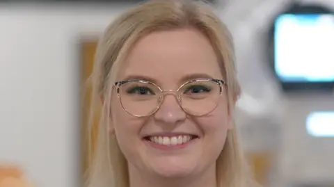 Jamie Niblock/BBC Ms Shuttlewood-Limb smiles at the camera. She has long blonde hair which has been partly tied back behind her head. She is also wearing glasses.