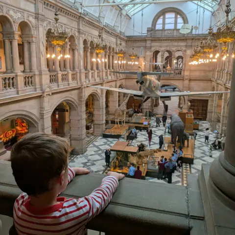 A child in a striped shirt leaning on a stone balcony inside a large museum hall with arched balconies, ornate lighting, a marble floor, and exhibits including a suspended aircraft, a giraffe model, and an elephant sculpture