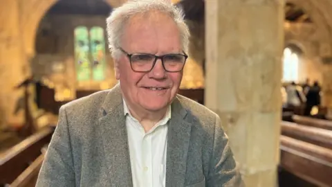 BBC/Seb Cheer An older man smiles at the camera. He is standing in a church with a few people standing in the background, among empty pews.