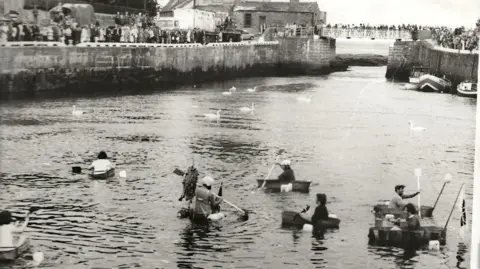 CADS A black and white photo of the first races, which shows several tin baths being paddled while spectators look on from the harbourside.