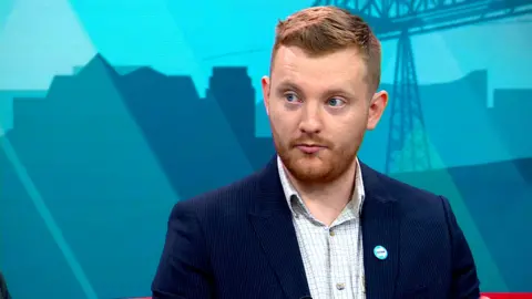 Chris Eynon sits with the teal backdrop of the BBC Politics North set behind him. He's wearing a navy pinstripe suit and checked shirt. He has short ginger hair and beard.