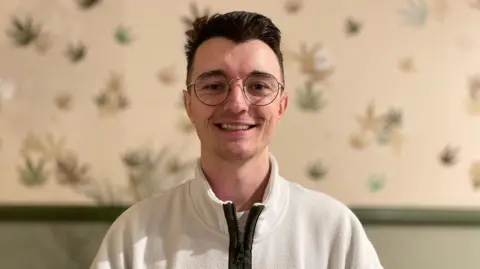 James O'Dell has short dark hair and wears round glasses. He's smiling and wearing a white fleece with a zip. He is standing in front of a wall with green panelling at the lower part, and different colours and sizes of cannabis leaves printed on the wall above.