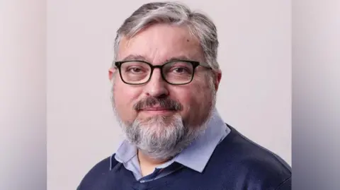 Gavin Forster Photography Councillor Ron Beadle. He has grey hair and a beard, and dark rimmed glasses.