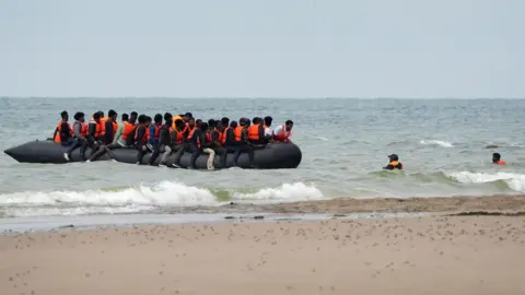PA Media A small, overcrowded inflatable boat floats near the shore, carrying numerous people wearing orange life jackets, believed to be migrants. Several individuals are in the water nearby, either swimming or wading toward the boat.