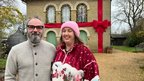 Tom Jackson/BBC Doug and Liz Pecarski are looking at the camera and standing on a large gravel drive in front of a brick house. SHe is wearing a Christmas top and he has a grey cardigan on. There is a large fabric ribbon and bow over the house and decorations in the windows.