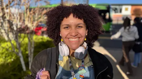 Student Kaitlyn smiling. She is wearing headphones around her neck, has yellow earrings on and a lanyard. 