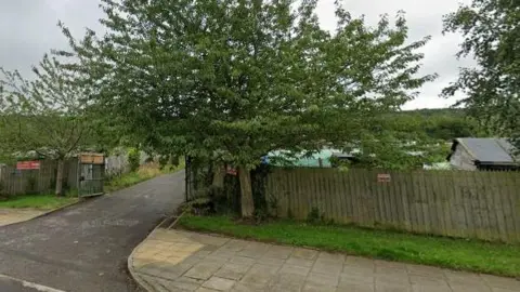 Google A wooden fence which lines a path and road. A large tree is at the entrance of an open wooden gate. An allotment can be seen behind the fence.