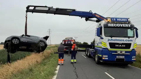 Cambridgeshire Police A silver and black car is being craned out of a river which is running on the left. Two people are standing in a road watching the car being winched.
