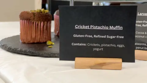 A small black sign detailing the ingredients of a Cricket Pistachio Muffin. The sign reads "Cricket Pistachio Muffin. Gluten-free, refined sugar-free. Contains: Crickets, pistachio, eggs, yoghurt"