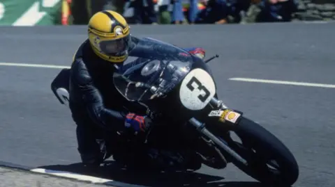 Joey Dunlop racing one of his bikes at the Isle of Man, it is black and has a white circle with the number 3 on it. He is wearing a black racing suit and a yellow helmet. He is coming around a corner on the bike as the image is taken. There are spectators in the background watching.