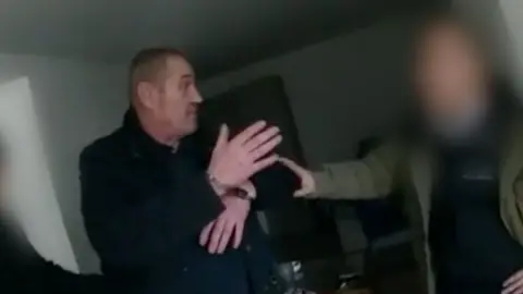 A man in handcuffs is being held by another person with a blurred face in a dimly lit room.
