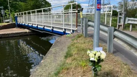 BBC A metal bridge over a canal in Exeter. A bouquet of white flowers and a card have been placed at the base of a pole next to the bridge.