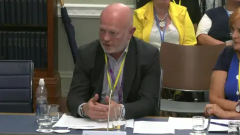 Chris Hagan is bald on top with grey hair at the sides and a grey beard. He is wearing a suit jacket and open-necked shirt with a yellow lanyard around his neck