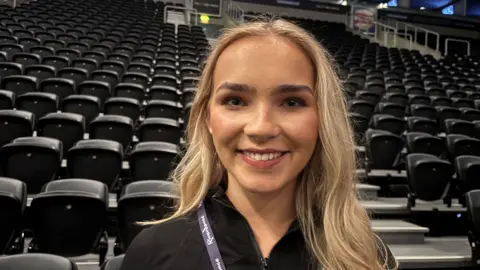 BBC Riverdance cast member Caitlyn is smiling at the camera, standing in front of the seating bank in the SSE arena. She is wearing a black jacket with her Riverdance Lanyard around her neck.