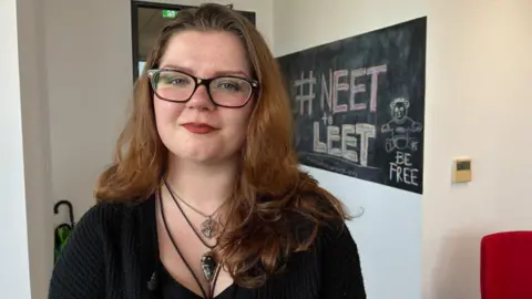 Amelia Wright has long, light brown hair and is wearing dark-framed glasses, a black top, black cardigan and two necklaces with large pendants. She is standing in front of a board which has #NEET to LEET written in chalk alongside the message BE FREE.