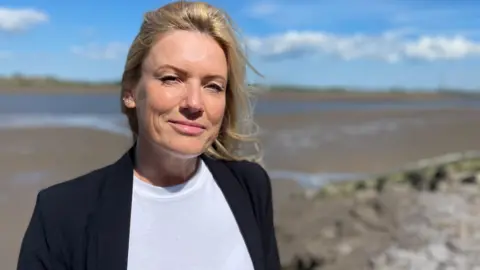 Rachel has long hair, is wearing a white t-shirt and a black jacket. She is standing on the banks of the river Wyre.