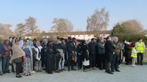 A crowd of around 40 people, wearing dark clothing, with some women in headscarves, looking quiet and sombre.