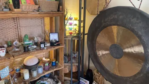A corner of a room is shown with a bookcase full of candles, a Buddha statue and crystals. Next to it on the right of the photo is a large gold and black gong.