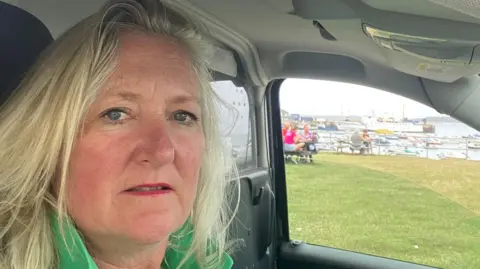 LDRS Sam sitting in a car with grass and a harbour in the background. She has long blonde hair, red lipstick and is wearing a green top.