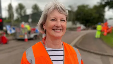 Liz Leffman has medium length grey hair and is smiling. She is wearing an orange hi-vis over a white and blue striped t-shirt. Behind her, contractors are fixing potholes.