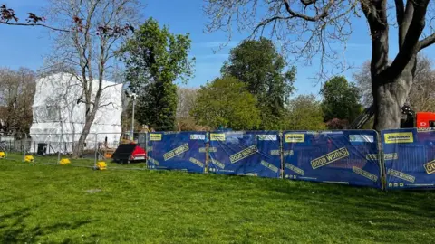 Alex Pope/BBC A large structure, covered in white covering, with scaffolding around it, a fenced off area, with blue covering up. A large area of grass in front of the fencing and trees all round it. 