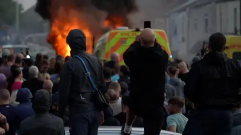 Three men dressed in all black look on at a crowd of people, a police van, and a large fire ahead of the van. 