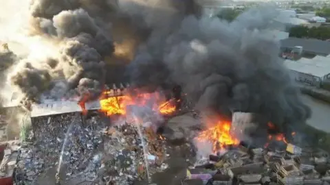 A fire at what looks like a scrapyard - with piles of burning rubbish and several buildings ablaze.