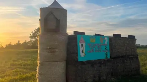 Comberton YFC A church made out of straw bales sitting in a grassy field against a sky as the sun sets off camera. There are round bales topped with a cone to form the church tower and rectangular bales form the body of the building. There is a blue sign on the sculpture saying The Cambridgeshire County Show Sat 30 May Abbots Ripton. 