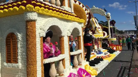 BBC People performing on a florally decorated float on a parade.