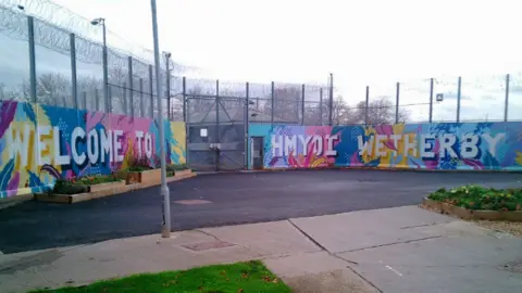The image shows the exterior of Wetherby Young Offender Institution with tall metal fencing topped with rolls of barbed or razor wire. A painted mural covers the perimeter wall, featuring colourful patterns and large white lettering that reads: "Welcome to HMYOI Wetherby". The ground is paved, with small planted flower beds in front of the mural.