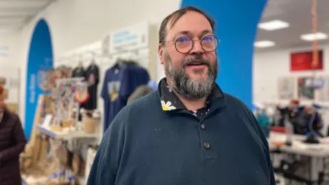 Tim Feake: a man with grey hair and beard and glasses, in a blue top with a flowery collar. He is in a charity shop and looking directly at the camera. 