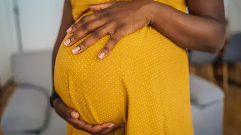 Close-up of pregnant black woman wearing a yellow dress with small white dots. The picture does not show the woman's face. She has both hands resting on her pregnant belly and she is standing in a room with a grey sofa behind her and some grey chairs and a table.