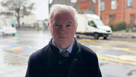 Man wearing a dark coat poses for a photo on a busy road