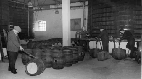 Royal William Yard Archive A black and white photograph of the brewery. There is a man in a suit and flat cap rolling a barrel along the floor with a stick. There are about a dozen wooden barrels standing on the floor. There are three men standing in a line wearing overalls and they are filling a barrel each from an enormous barrel.