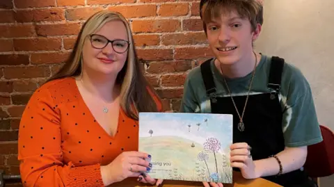 Supplied A woman in an orange top holding a book up to the camera. A man is on her left wearing a dark hat, green t-shirt and black dungarees. 