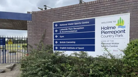 BBC The entrance to Holme Pierrepont Country Park - there is a blue and white sign affixed to a brown brick wall, on the left side of the wall is a blue gate/ A bush is visible in the background