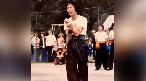 Liverpool Hung Gar Kung Fu Club Image from the 1980s of Master Jimmy Chan with black hair wearing a martial arts outfit including a white t-shirt with red Chinese writing on it and long black satin trousers and black studded belt and wristbands. He is holding a martial arts pose with his arms crossing showing his muscles in a park. People are watching him while students watch on. 