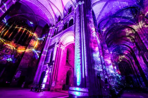 Peter Walker A galaxy illumination inside the cathedral. There is a bright yellow circle on the left against a backdrop of pink and purple. On the right the columns and the ceiling are lit up in a mix of blue, yellow, and pink in swirly shapes.