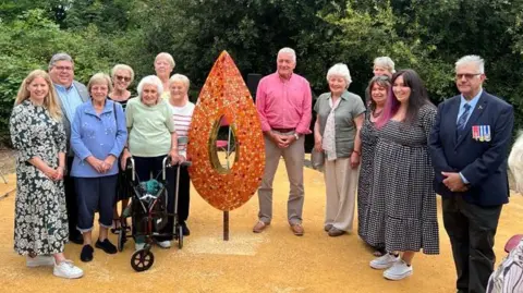 An orange, red and yellow leaf sculpture on a light hardcore surface. The sculpture is about the same height as the men and women surrounding it, all smiling towards the camera.