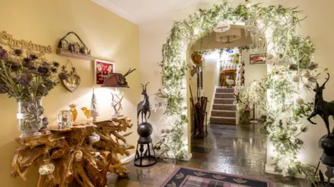 Kirsty Anderson Festively decorated hallway with a rustic wooden console table, Christmas ornaments, and a large archway adorned with greenery, white flowers, and twinkling lights leading to a staircase.