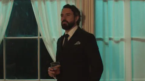 WORLDPRODUCTIONS/PA Tom wears a black suit with a white shirt and red tie, He has a grey pocket square. He holds a glass tumbler