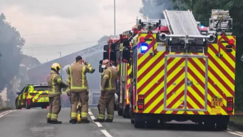 A row of fire engines and four firefighters in the foreground, on a road. In the distance is a fire car.